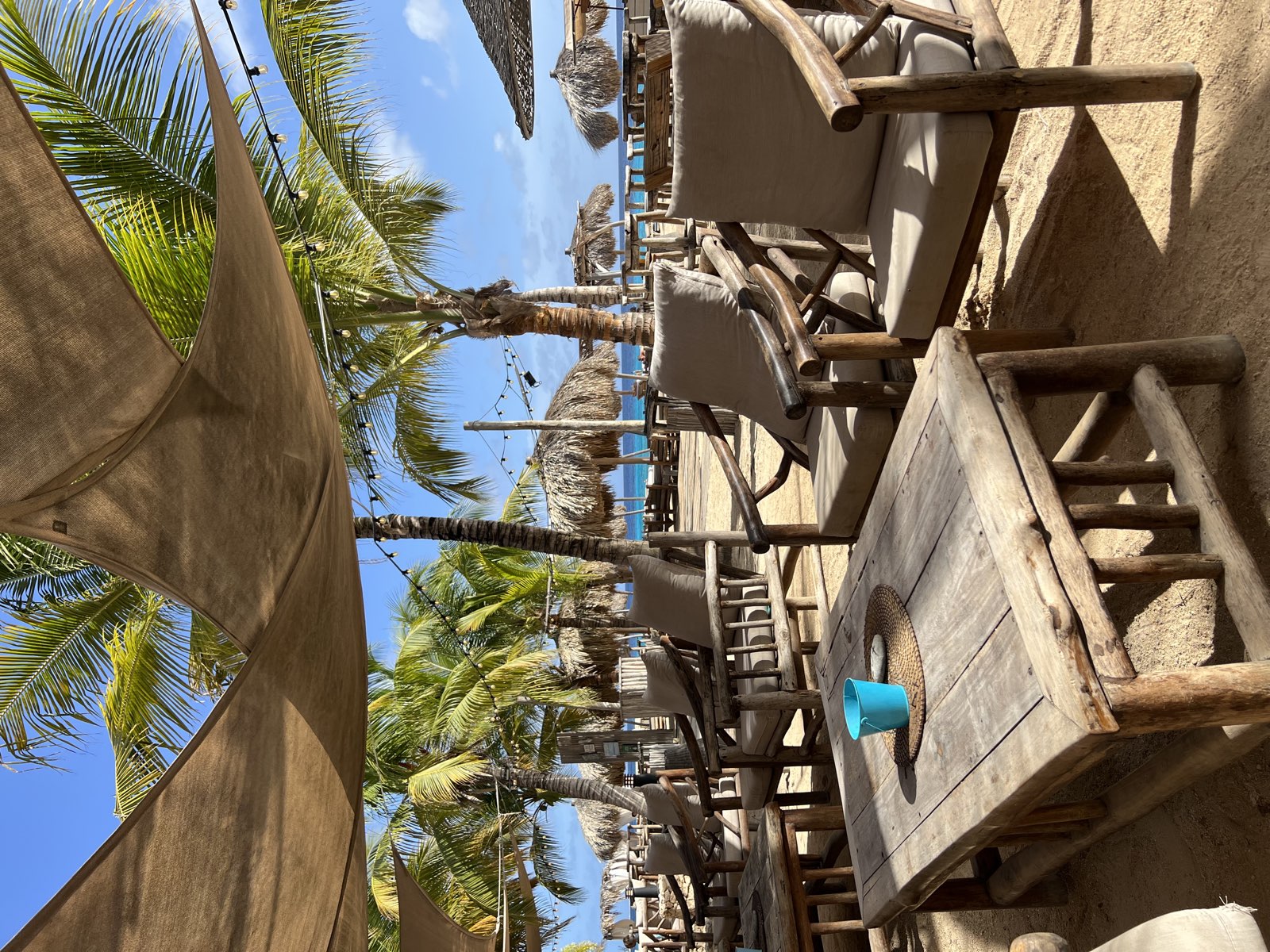 Wooden lounge chairs and a table on a sandy beach beneath shade sails, with palm trees and the ocean beyond.